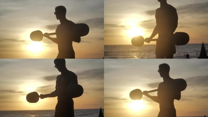 年轻的肌肉发达的人在日落背景下在海滩上做杠铃提升。运动员训练二头肌。