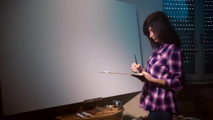 集中的女艺术家在黑暗照明艺术工作室用画笔在木制调色板上混合丙烯酸色。自信的画家为留在木制画架、当代艺