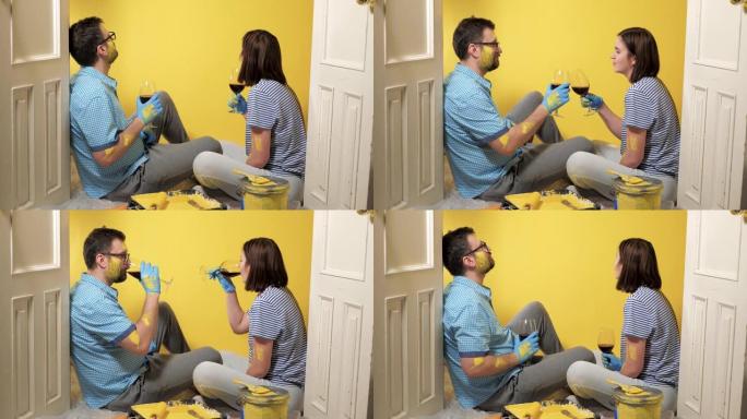 疲倦的男女夫妇坐在地板上，看着涂着的黄色墙壁和喝酒，他们累了但很高兴。油漆修复工作超过了概念。慢动作