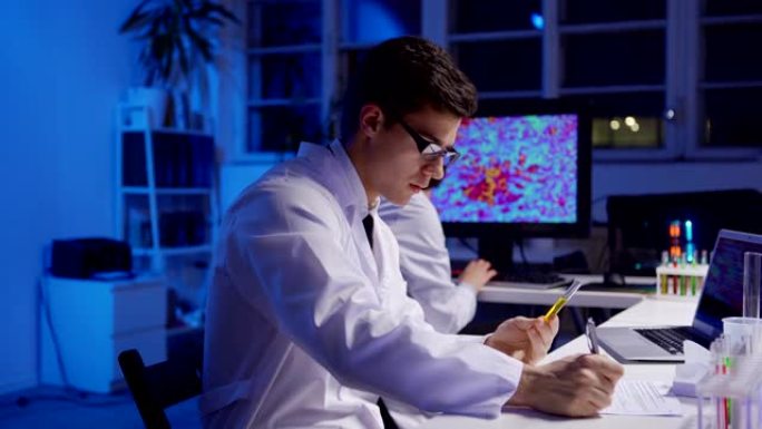 使用笔记本电脑跟踪年轻男性科学家的镜头侧视图，检查试管中的液体并写下测试结果，而他的女同事在实验室中