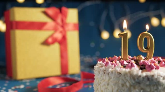 白色生日蛋糕编号19点灯燃烧的金色蜡烛，蓝色背景带灯和用红丝带绑起来的礼物黄色盒子。特写