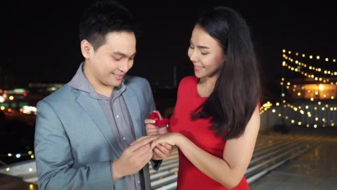 高清慢动作快乐的亚洲情人男人通过给她漂亮的结婚戒指和在浪漫之夜求婚来表达惊讶的红色连衣裙美女。浪漫求