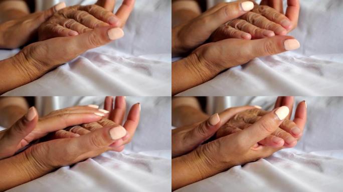 女人牵着老母亲的手躺在床上，表现出关怀或爱心。女儿在医院安慰年迈的妈妈皱巴巴的手臂。孙女温柔地抚摸奶