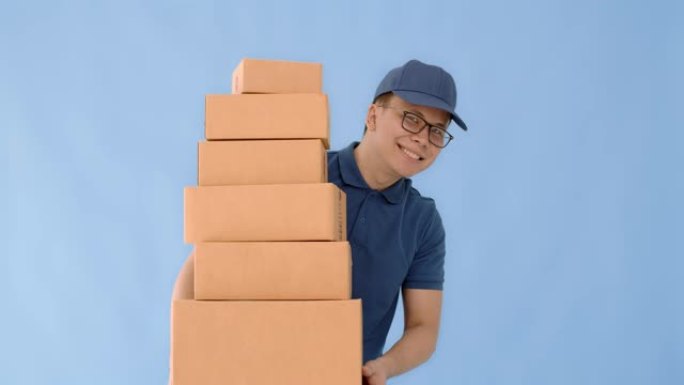 亚洲快乐送货员戴着一顶帽子，穿着一件蓝色衬衫，拿着纸质包装箱，孤立在蓝色背景上，面带微笑。邮政递送服