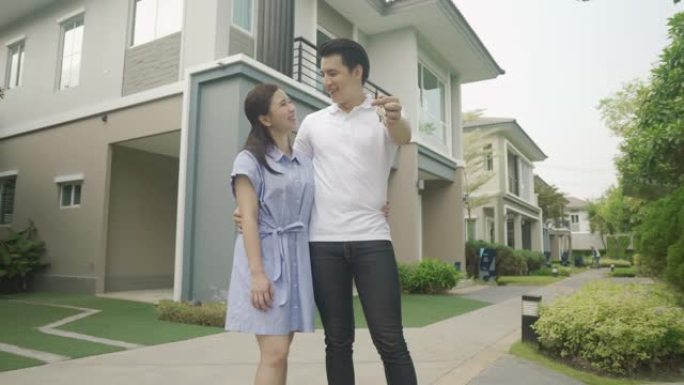 亚洲年轻夫妇站在一起拥抱在一起，拿着房子钥匙在他们的新房子前看起来很开心，开始新的生活。家庭、年龄、