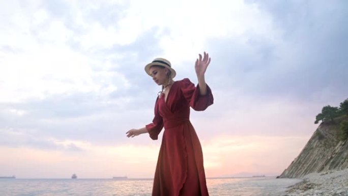 一个迷人的年轻女孩，穿着红色的夏装和草帽，在海浪和日落的背景下，小心翼翼地爬行在岩石海岸的石头上。