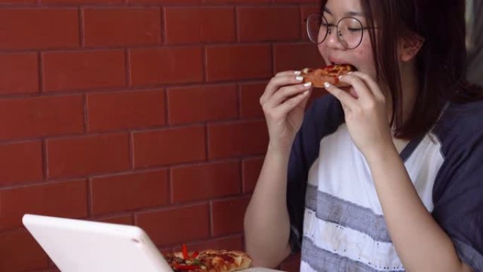 年轻的亚洲女性从饮食中休息一下，选择披萨作为餐食，并从数字平板电脑中寻找媒体