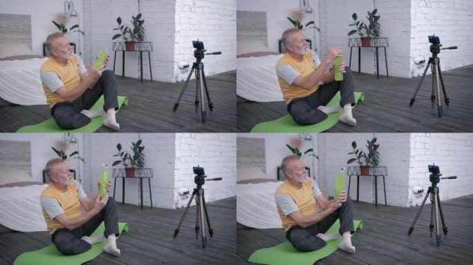 有影响力的老年男性博客作者在智能手机上记录了维生素水的视频评论，推荐他的订阅者坐在美丽的室内瑜伽垫上