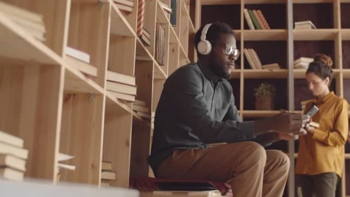 年轻的黑人在书店里戴上耳机并使用智能手机