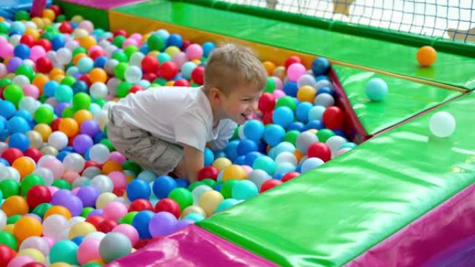 金发小男孩在不同的侧面散球，躺在游戏中心大干划水池中的多色塑料球上。对着镜头微笑。在游戏室玩得开心。