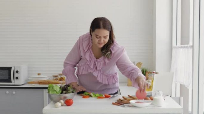 胖女人正在准备沙拉，西红柿，火腿和汉堡包，以制作汉堡包，同时在厨房的笔记本电脑上观看如何在线制作汉堡