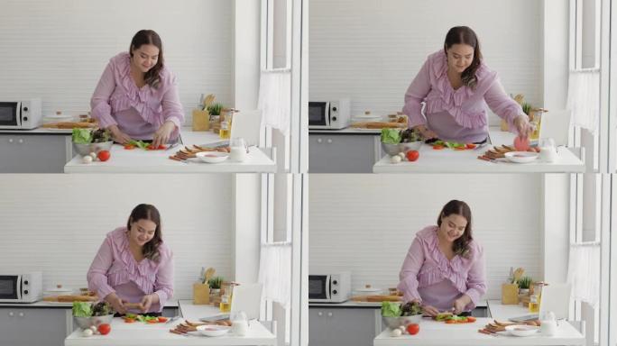 胖女人正在准备沙拉，西红柿，火腿和汉堡包，以制作汉堡包，同时在厨房的笔记本电脑上观看如何在线制作汉堡
