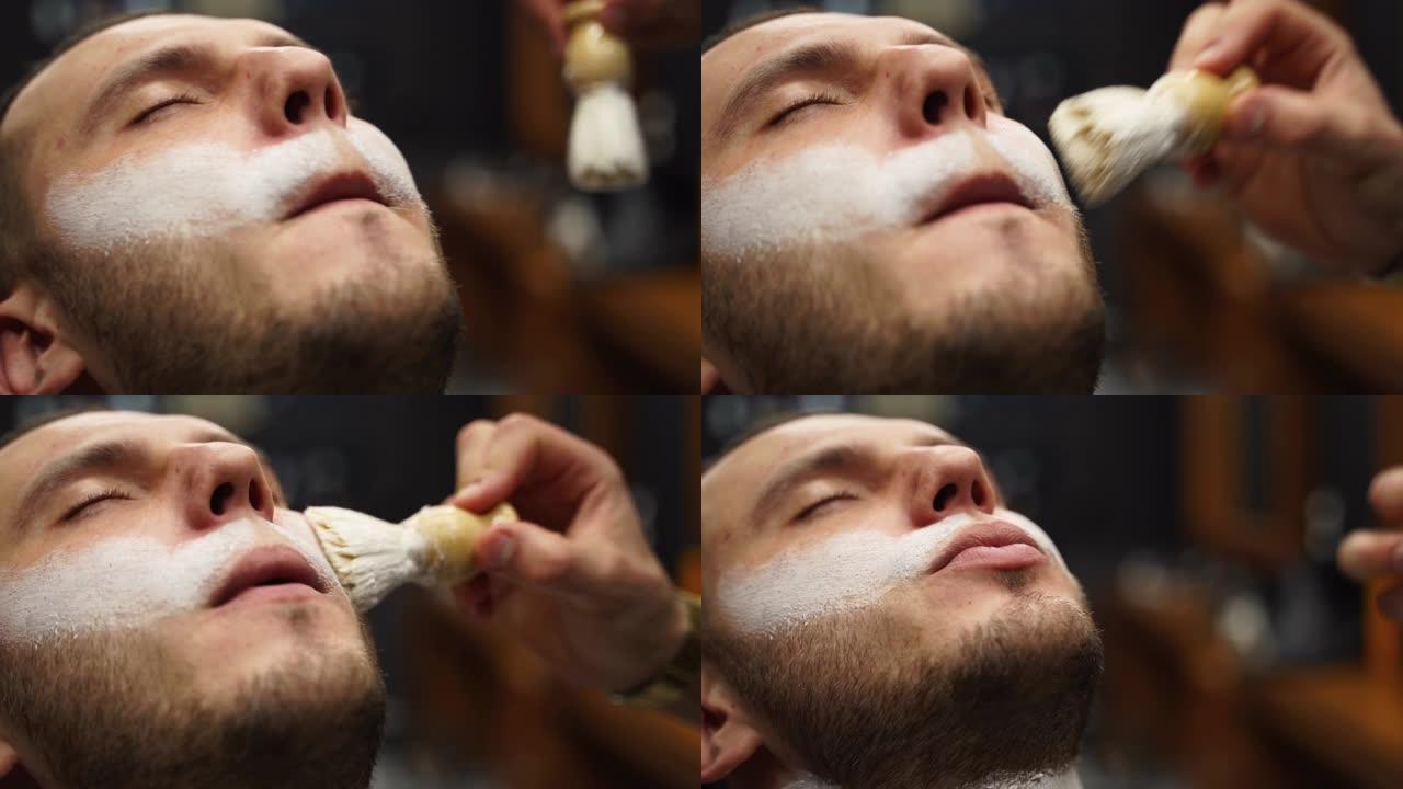 理发师用剃须刷工作。男人在理发店用剃须膏起泡的特写镜头。在老式美发工作室刮胡子前，客户脸上有白色泡沫