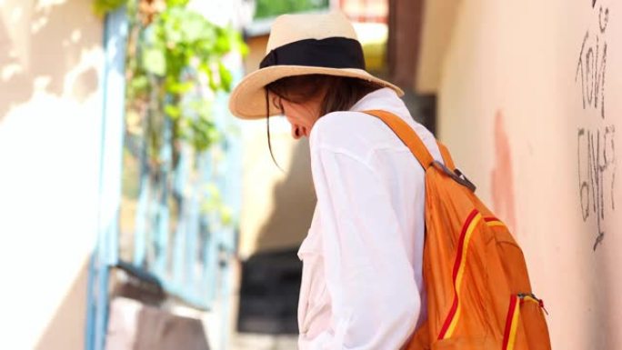 穿着草帽、牛仔裤短裤、白衬衫和橙色背包的年轻优雅女性游客享受狭窄街道