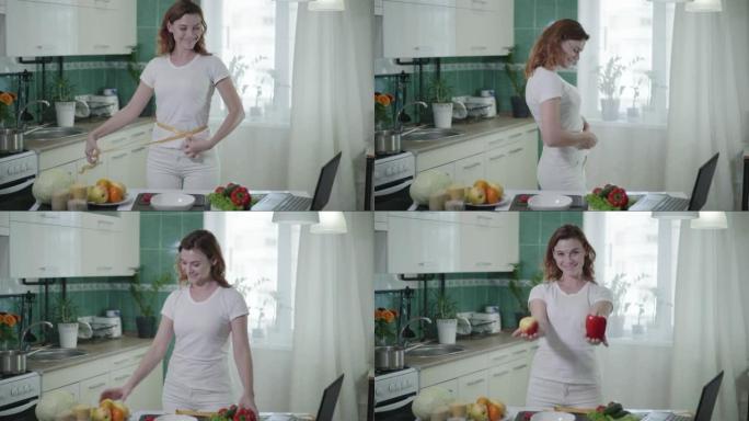 成功节食后的女孩肖像用卷尺测量她的腰部，手里拿着苹果和胡椒在厨房摆姿势