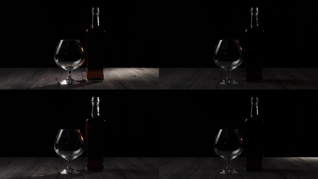 豪华白兰地。方形瓶子中的金色干邑白兰地站在黑色背景下的木制桌子上的玻璃杯附近。光线照在桌子上，玻璃在