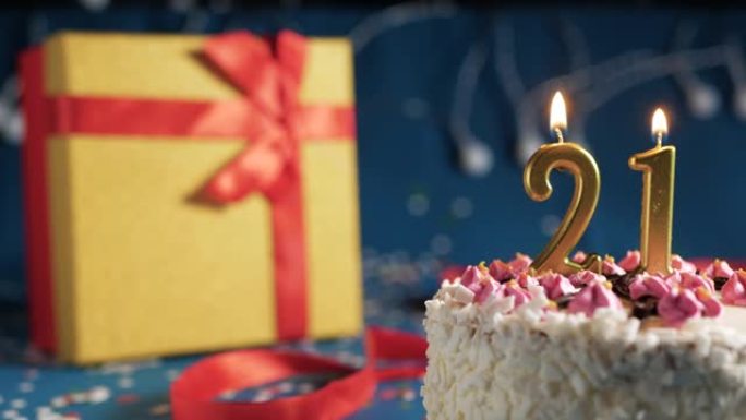 白色生日蛋糕编号21点灯燃烧的金色蜡烛，蓝色背景带灯和用红丝带绑起来的礼物黄色盒子。特写