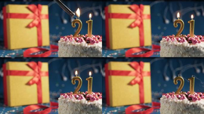 白色生日蛋糕编号21点灯燃烧的金色蜡烛，蓝色背景带灯和用红丝带绑起来的礼物黄色盒子。特写