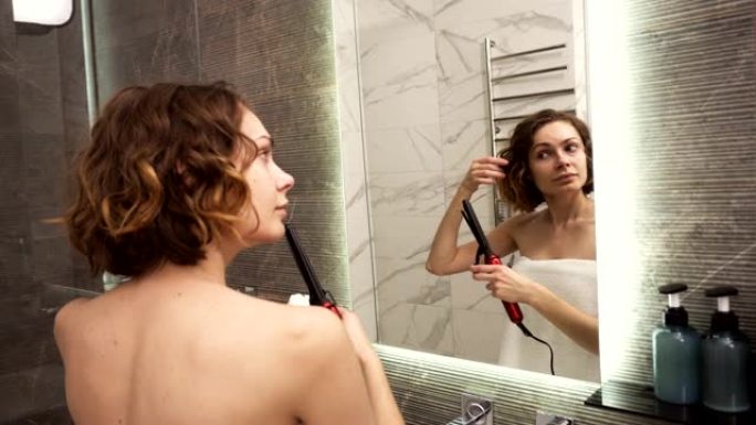 一个穿着白色大浴巾的年轻女子在镜子前用卷发棒烫头发。一个女孩洗完澡后在浴室里做发型。透过镜子看