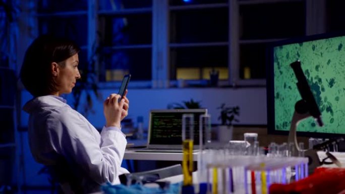坐在实验室桌子上的手机上的实验室外套女科学家的侧视图跟踪照片，放大了她电脑屏幕上的微生物图像
