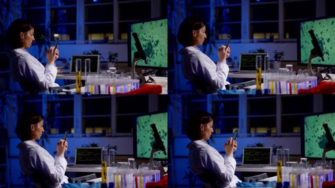 坐在实验室桌子上的手机上的实验室外套女科学家的侧视图跟踪照片，放大了她电脑屏幕上的微生物图像