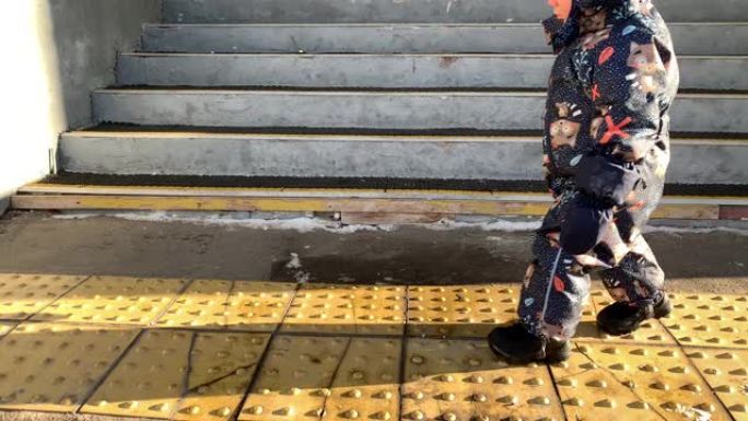 一个穿着冬季连身裤的小孩在黄色的触觉瓷砖上行走，供视障人士在冬日的阳光下行走。俄罗斯的冬天。