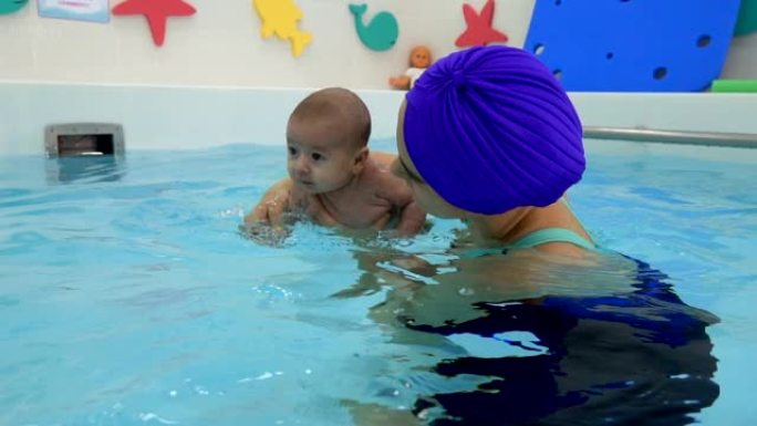 一位快乐的母亲正在儿童游泳池的水中与新生婴儿一起游泳。她用手支撑婴儿，拥抱他并亲吻他。婴儿在儿童游泳