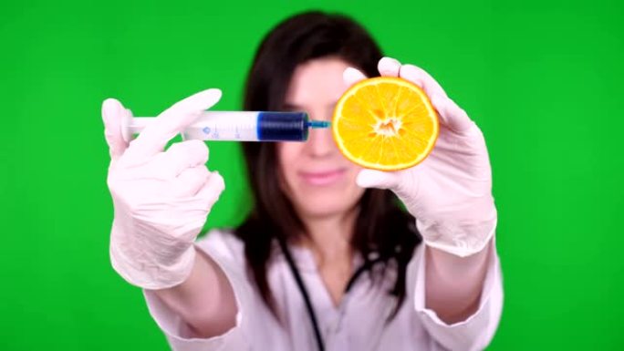 年轻的女医生，营养师身着白色医用制服，用听诊器，用注射器将一些蓝色液体注入一半的橙色。绿色背景