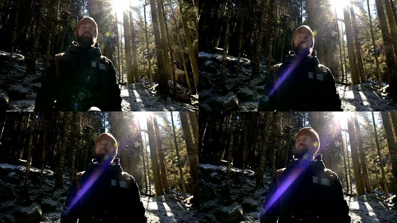 一位认真的大胡子摄影师的肖像，背着背包和相机站在针叶树的冬季森林中，在背光和阳光照射下