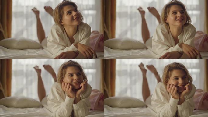 微笑的高加索女孩躺在柔软的床上抬头的肖像。积极的年轻女子穿着白衬衫摇晃双腿。生活方式，幸福，休闲。