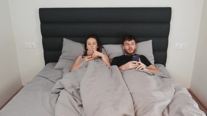 女性和男性躺在卧室里，看着他们的智能手机。关系危机。关系困难。年轻的已婚夫妇在床上看着智能手机并在互