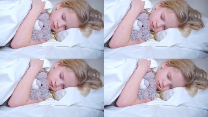 熟睡的小女孩在梦里拥抱一只泰迪熊。漂亮的金发孩子一大早就睡在床上，穿着白色床上用品。4k镜头