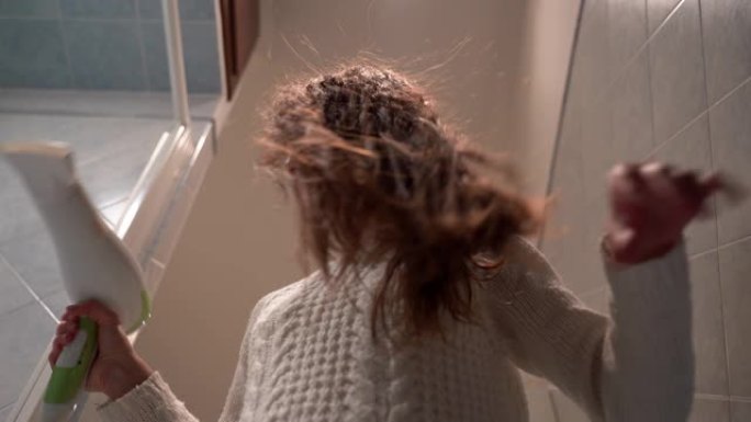 穿着白色毛衣的迷人年轻女孩在家用现代吹风机吹干她美丽的棕色长发。黑发女孩在早上洗澡、美容和日常生活后