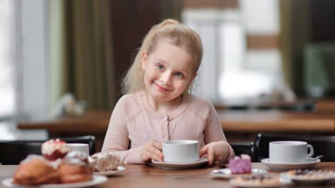 快乐微笑的女婴坐在桌旁端着一杯茶摆姿势。在4k红色相机上近距离拍摄