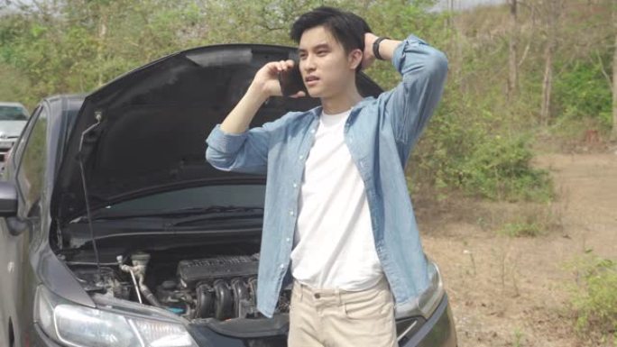 亚洲年轻的不快乐男子在乡村公路上开着的引擎盖抛锚的汽车前用手机打电话寻求帮助。旅途中的破车在路上。