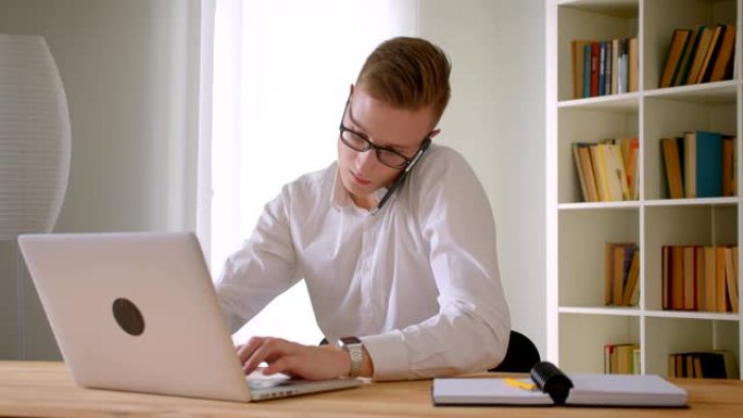 戴眼镜的年轻英俊的高加索商人的特写肖像在办公室的笔记本电脑上打电话