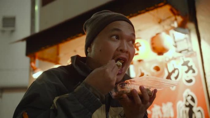 亚洲旅游男子晚上在城市街道吃日本食物章鱼烧。