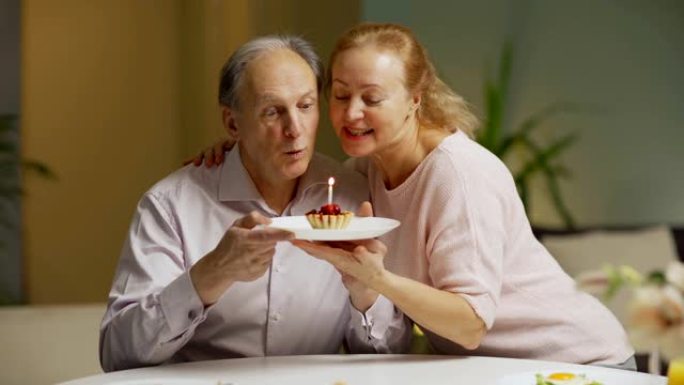 慈爱的妻子给坐在早餐桌上的丈夫蜡烛蛋糕。快乐的高级男人在蛋糕上吹蜡烛，得到妻子的祝福和亲吻