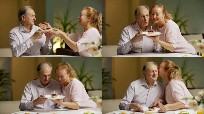慈爱的妻子给坐在早餐桌上的丈夫蜡烛蛋糕。快乐的高级男人在蛋糕上吹蜡烛，得到妻子的祝福和亲吻