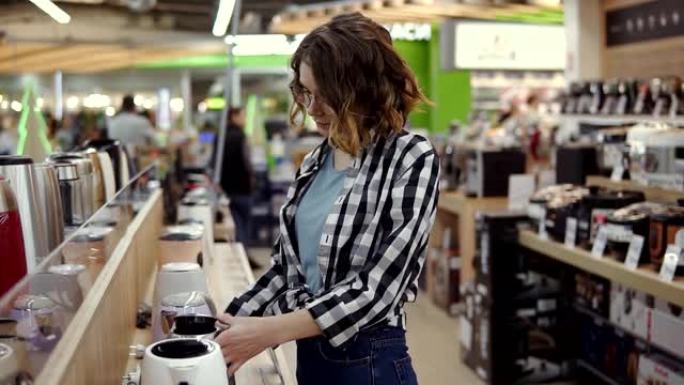 侧面的一个年轻愉快积极有吸引力的女人在家用电器商店选择电子水壶，站在一排，检查水壶