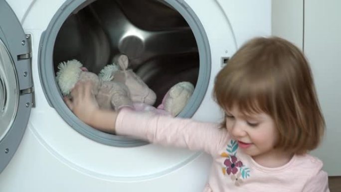 小高加索3岁女孩的特写镜头坐在地板上，把洗衣机的毛绒玩具洗。儿童清洗玩具，关闭舱门并打开家用电器。