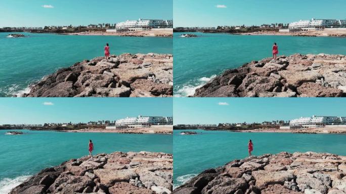 岩石海滩悬崖上的红衣女子。穿着红色连衣裙的女士在塞浦路斯珊瑚湾享受阳光和暖风。穿着红色和白色连衣裙的