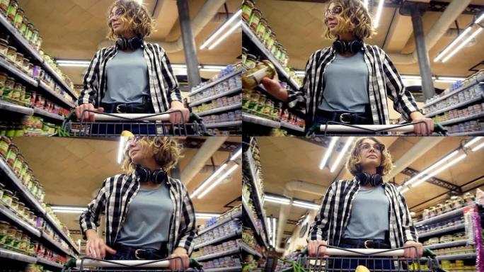 穿着格子衬衫和脖子上的耳机的开朗女人正走在杂货店里，转向里面装有食物的购物车，环顾四周装有产品的架子