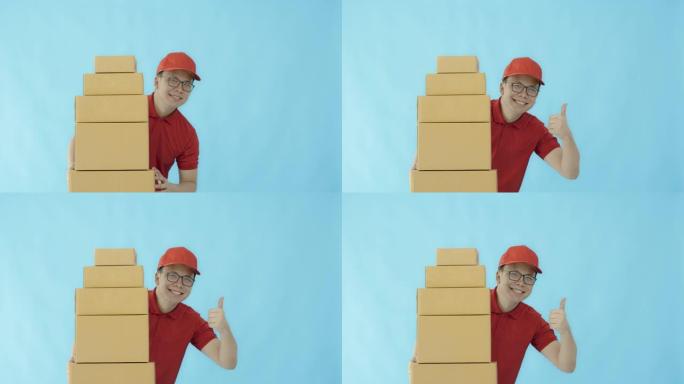 亚洲快乐的送货员戴着一顶帽子，穿着一件红色衬衫，拿着纸质包装箱，孤立在蓝色背景上，面带微笑。邮政递送