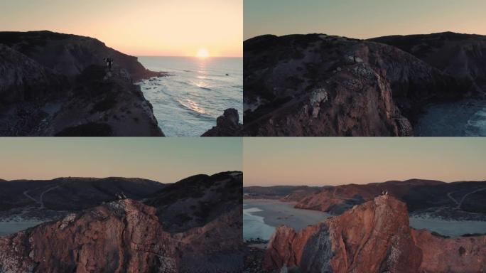 女子和男子冲浪者站在悬崖/岩石的边缘，在日出/日落时观看海浪的惊人航拍。