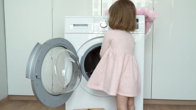 小微笑可爱的3岁女孩的肖像放入洗衣机玩具进行洗涤，关闭舱门并打开家用电器。儿童洗涤玩具