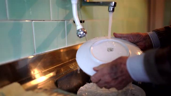 老人的手在家里的水龙头下洗碗。退休老人在做家务，养老金领取者在厨房打扫脏盘子。老人的日常生活