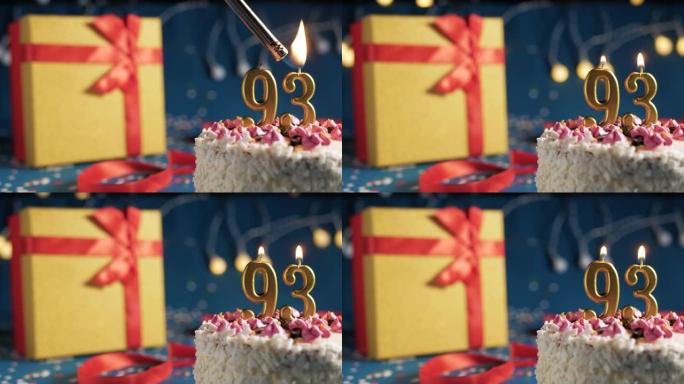 白色生日蛋糕93号点火器燃烧的金色蜡烛，蓝色背景带灯和用红丝带绑起来的礼物黄色盒子。特写