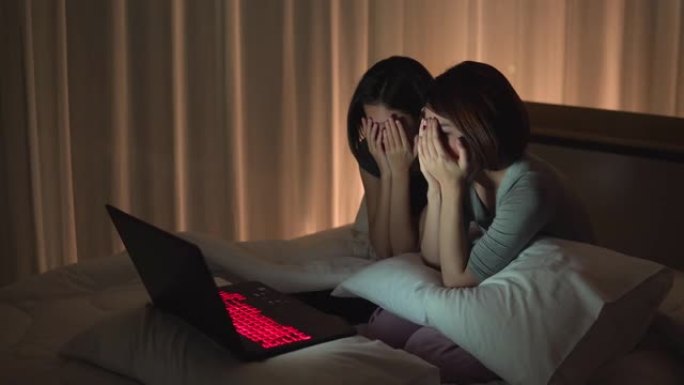 侧视图: 两个年轻的女性朋友在晚上通过看笔记本的恐怖系列在床上一起分享片刻