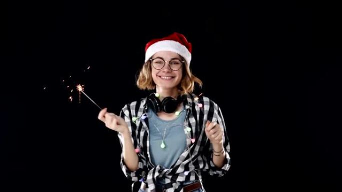 一个可爱的女人戴着圣诞老人的帽子，五颜六色的花环和脖子上的耳机，两个烟火在黑色背景的圣诞晚会上跳舞。
